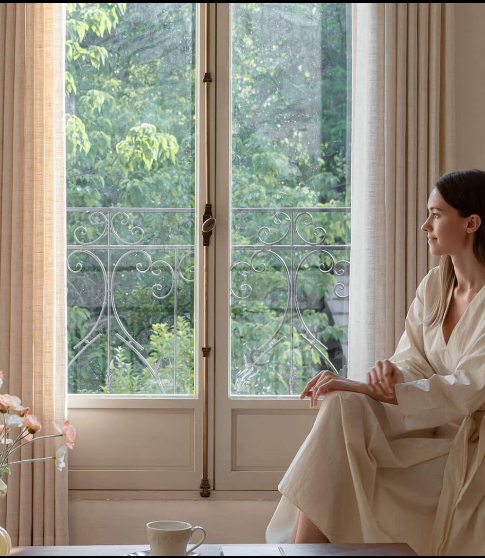 Woman in cream silk robe seated by a Parisian window overlooking lush greenery — embodying the calm intentionality of a modern skincare ritual