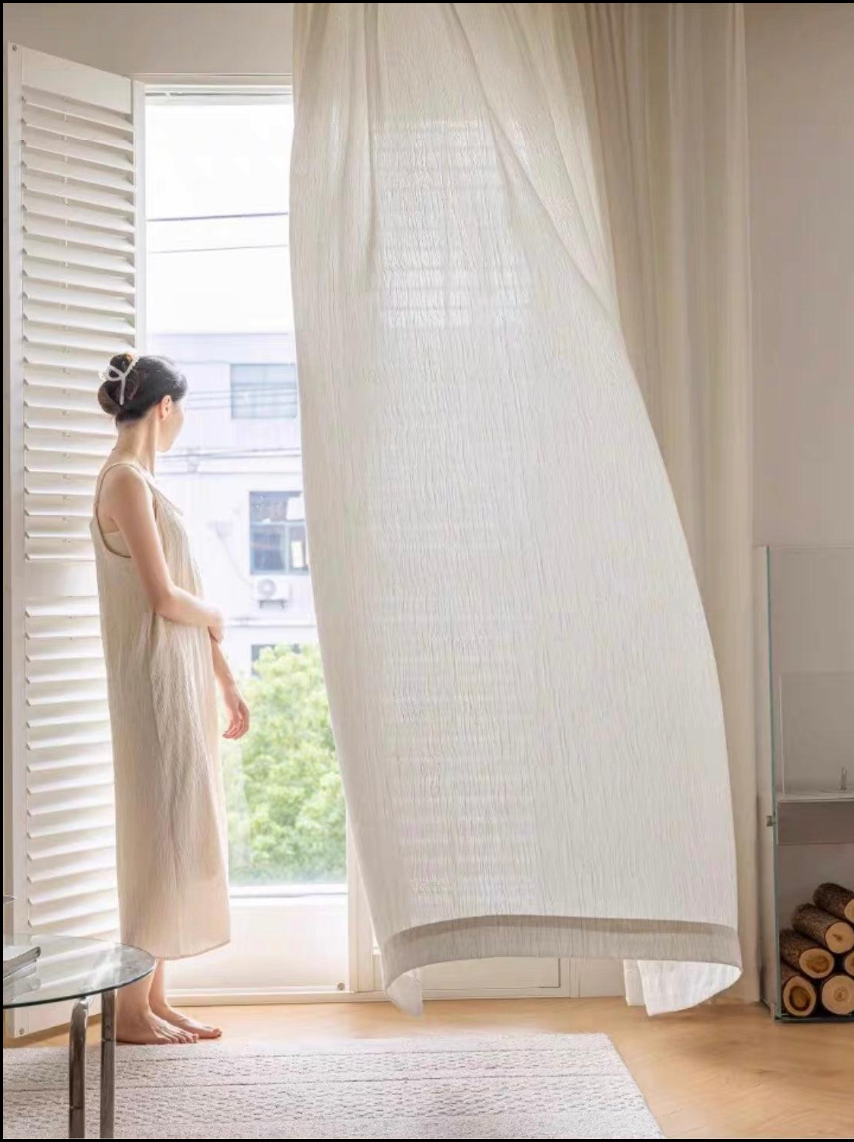 Woman in cream linen dress standing barefoot by a billowing sheer curtain and open window — a moment of stillness and intentional morning ritual